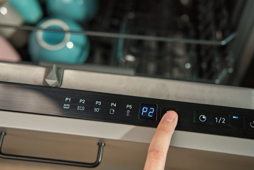 A finger on the top-edge control strip of a dishwasher, P2 lit on the small display beside the indicator lamps