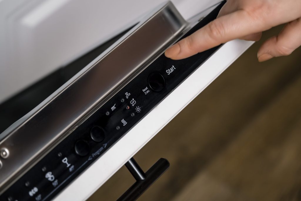 A finger on the start button of the control strip along the top edge of an open dishwasher door