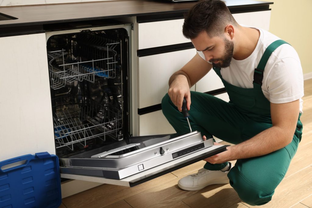 A technician in green overalls kneeling at an open built-in dishwasher, working on the door with a screwdriver