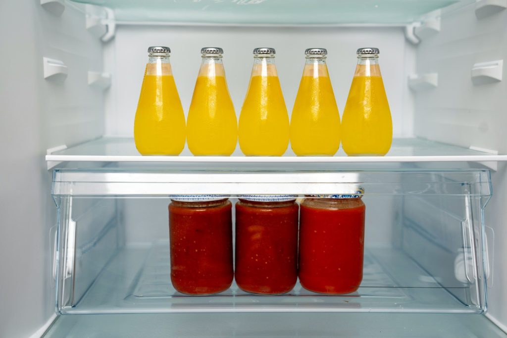 Five bottles of juice on a glass shelf above three jars of sauce