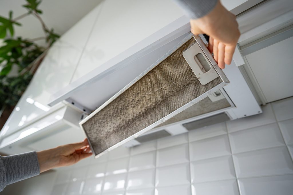 Two hands lifting a grease-caked mesh filter out of a slide-out cooker hood