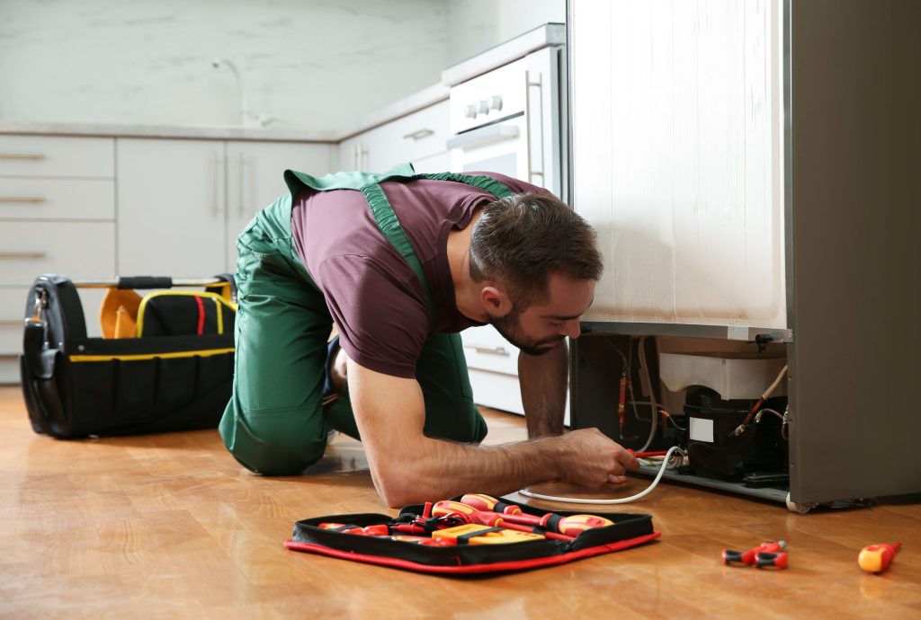 A technician working behind a refrigerator, tool roll on the floor