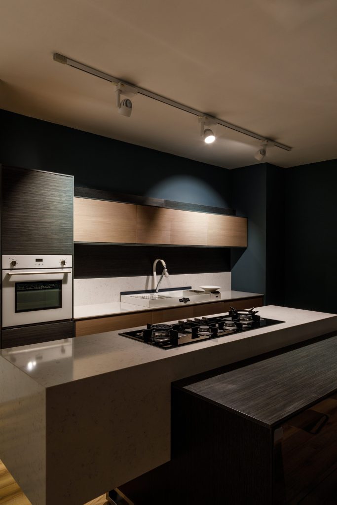 A dark blue kitchen at night, a gas hob set into a pale stone island, a white oven column at the left