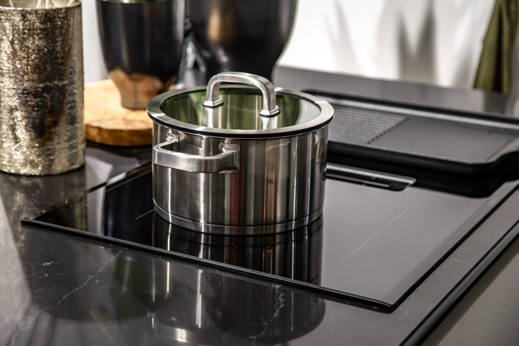 A steel pan standing on a black glass cooktop in a dark kitchen