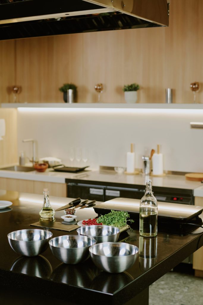 A black island laid for prep — steel bowls, knives, herbs, a bottle — under a stainless island hood, wood wall behind