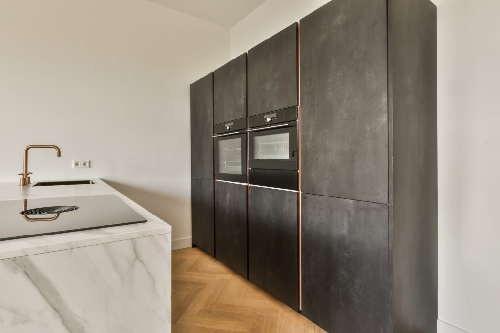 A dark tall cabinet run with two built-in ovens, copper reveals, marble island