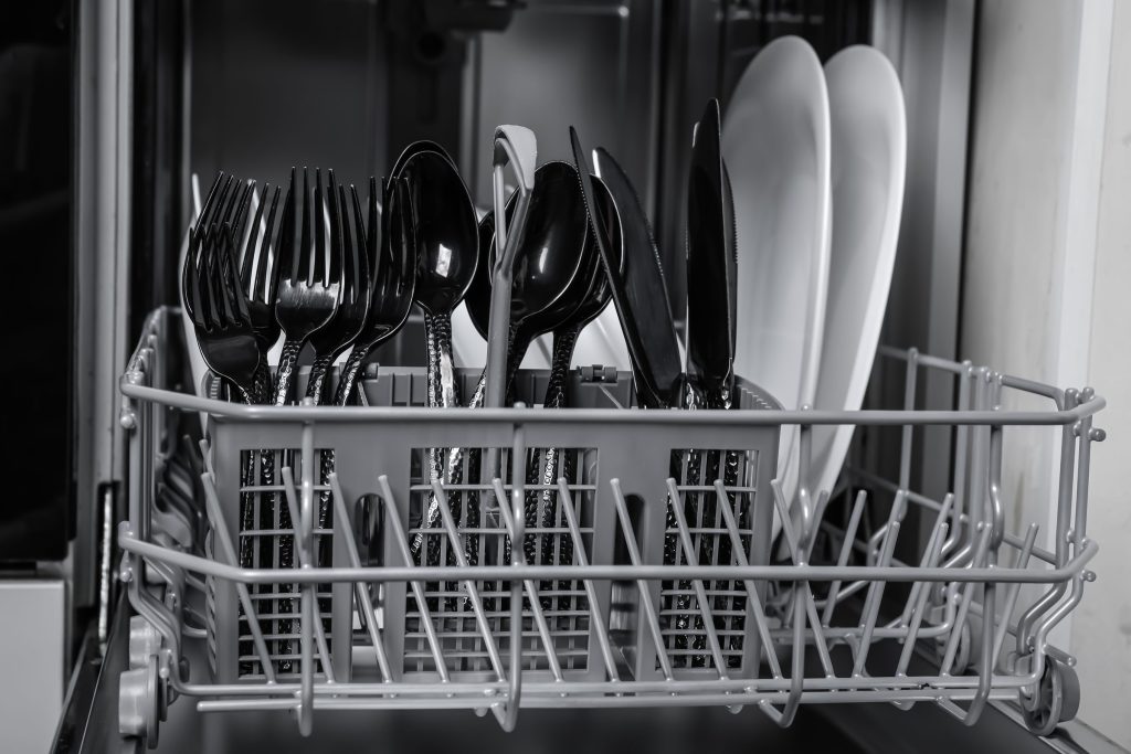 The cutlery basket of an open dishwasher, forks and spoons upright, plates behind, in near monochrome