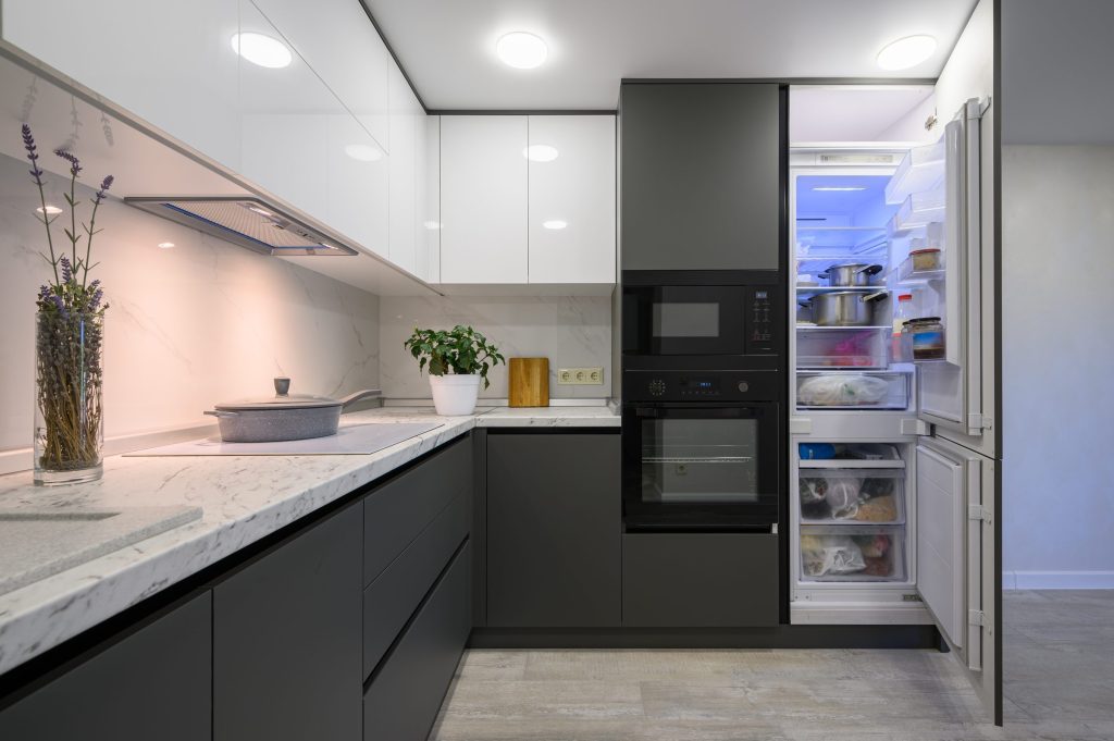 A grey and white kitchen with an oven and microwave stack, and a refrigerator standing open at the end of the run