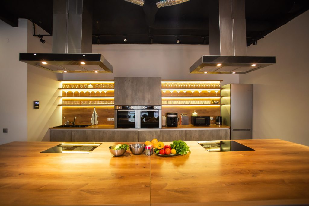 A wide oak island with two induction hobs under two stainless island hoods, glassware lit on shelves behind