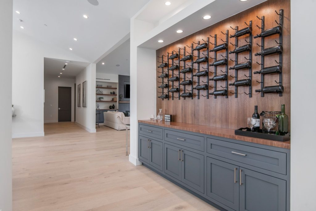 A wall of bottles on black steel racks over a grey cabinet run, in a bright hallway
