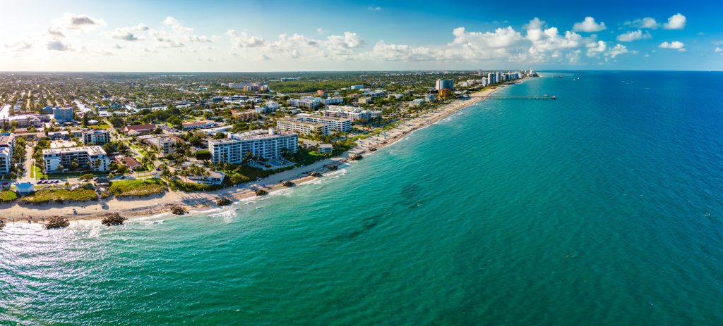 An aerial view along a South Florida coastline, low-rise blocks between the beach and the road