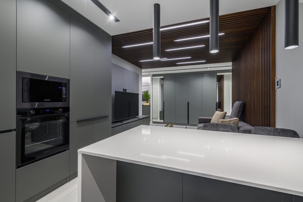 A grey kitchen run with stacked wall ovens opening onto a living room