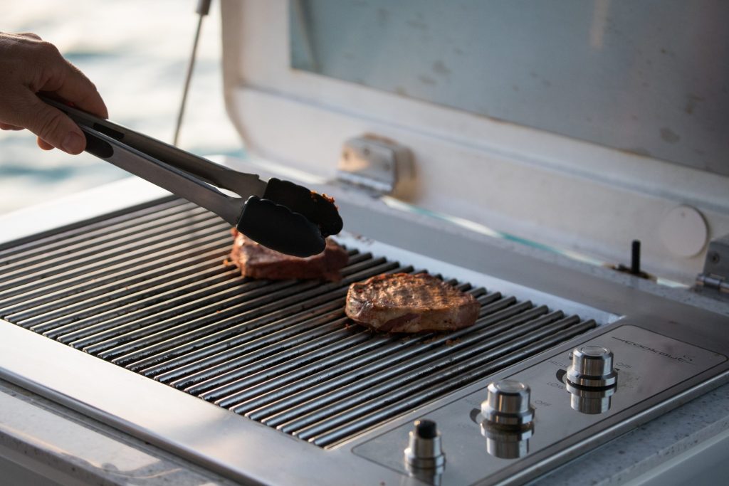 Tongs turning steaks on the grates of a stainless outdoor grill