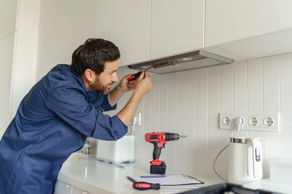 A technician in overalls working on the underside of a kitchen extractor hood with a screwdriver