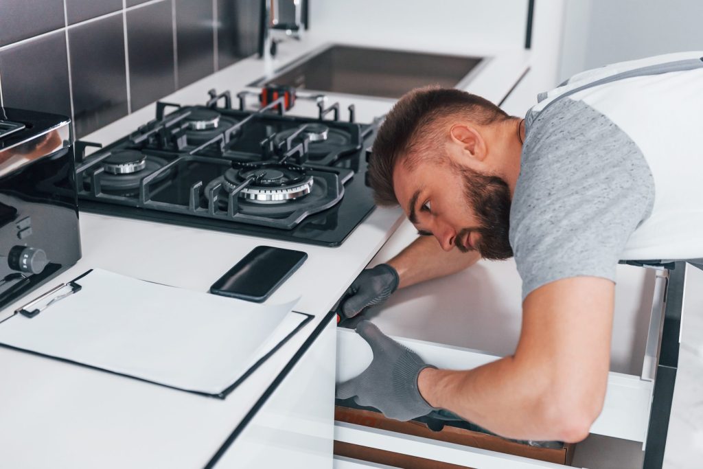 A technician leaning under a gas cooktop to check the cabinet below it, notes on the worktop