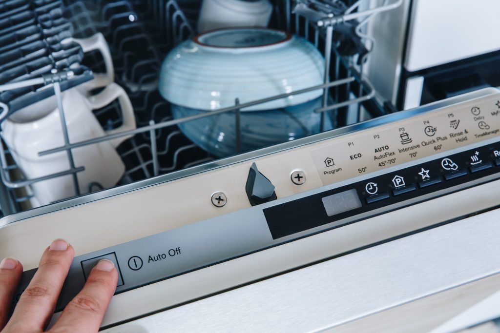 A hand at the control strip of an open dishwasher, the cycle buttons in a row along the door edge