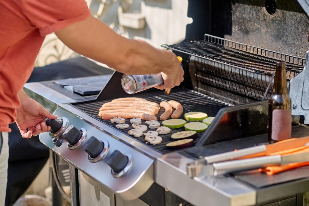 A hand at the control knobs of a stainless outdoor grill, food on the grates behind