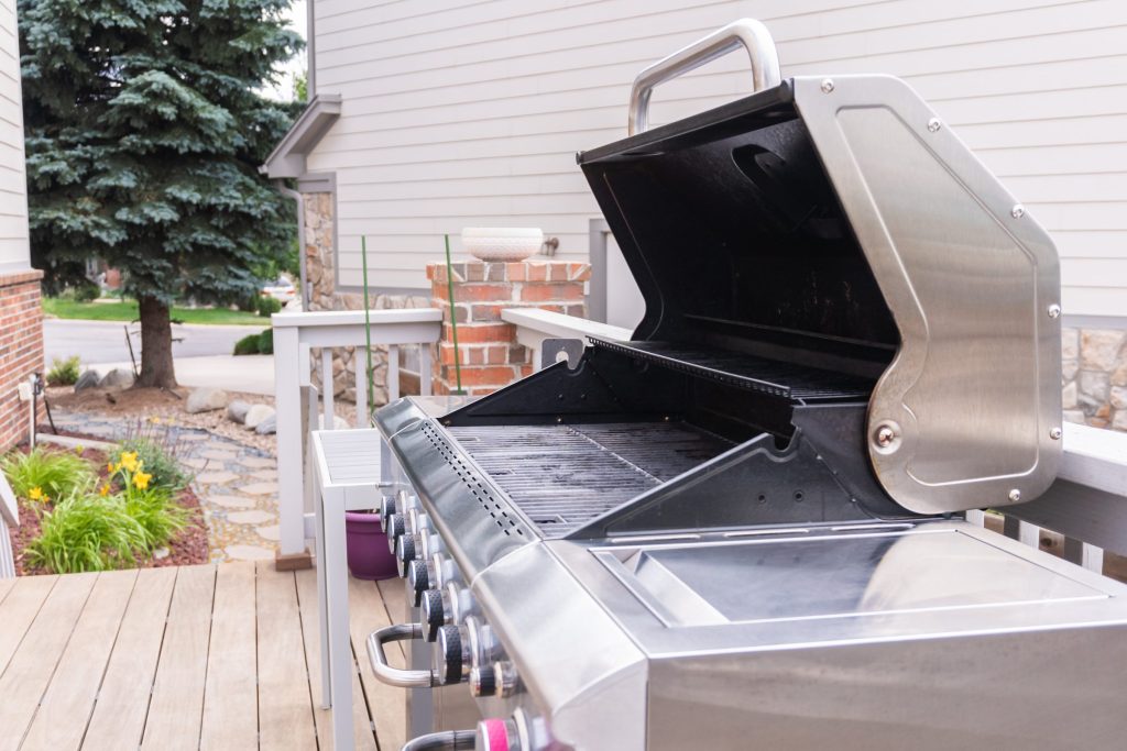 A stainless outdoor gas grill on a deck with the lid raised — burner tubes, grates and the row of control knobs along the front
