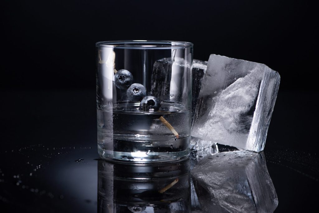 A glass of water with a large clear ice cube beside it on a dark surface
