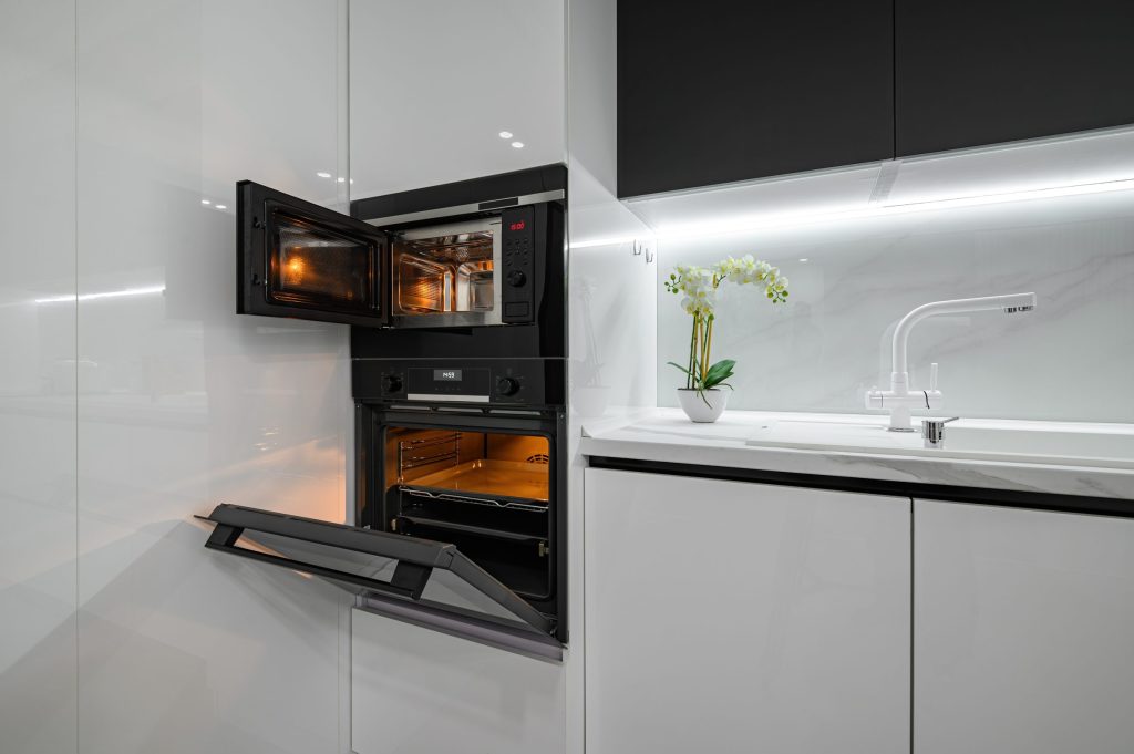 A built-in microwave standing open above a lit wall oven in a black and white kitchen
