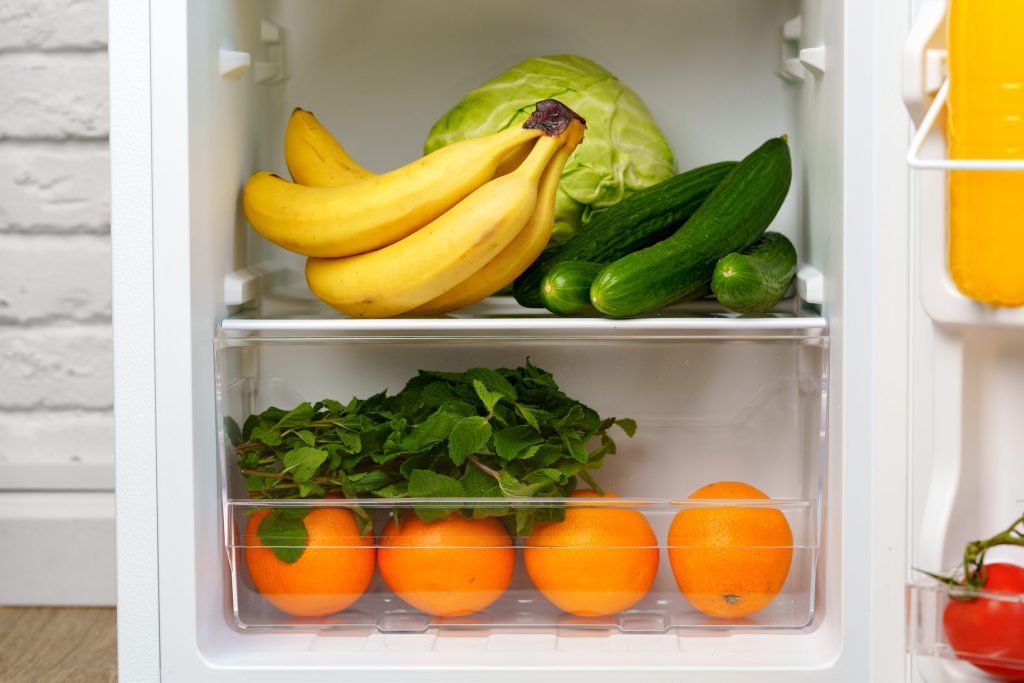 The inside of a refrigerator with fruit and vegetables on glass shelves and door bins alongside