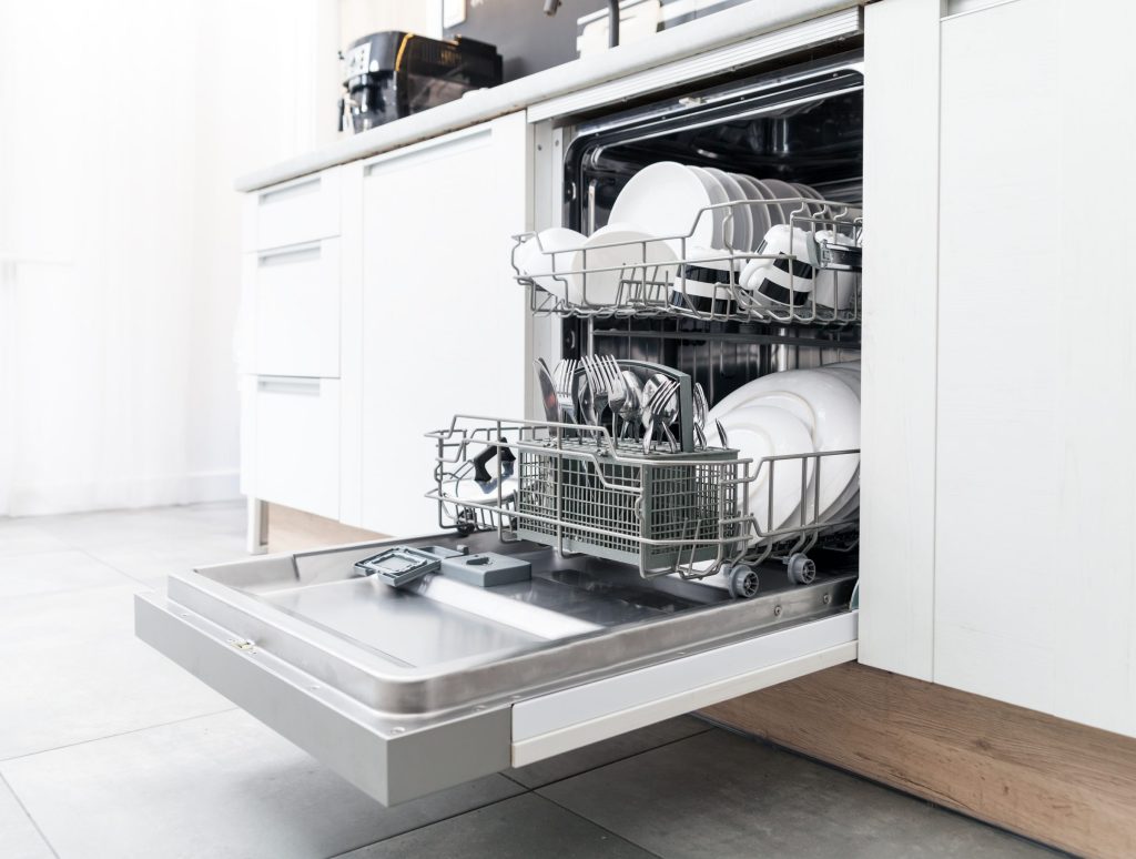 A dishwasher standing open in a white fitted kitchen, both racks loaded with clean china